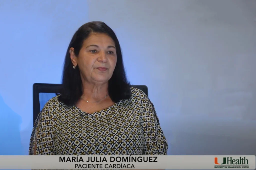 A woman with shoulder-length dark hair sits in front of a blue and white background. Her face is intentionally blurred. She wears a patterned blouse. Text at the bottom reads 'MARÍA JULIA DOMÍNGUEZ PACIENTE CARDÍACA' alongside the UHealth, University of Miami Health System logo