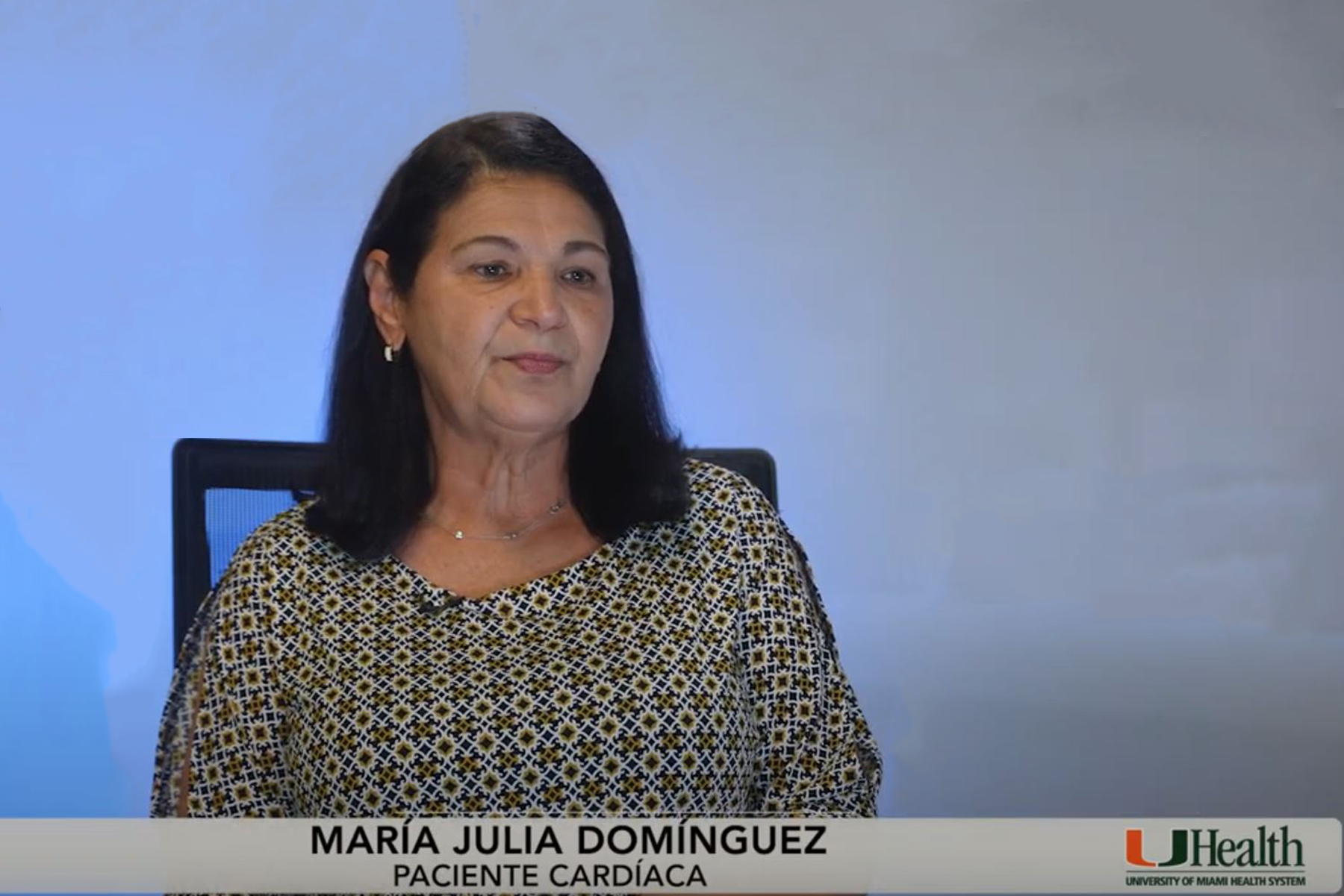 A woman with shoulder-length dark hair sits in front of a blue and white background. Her face is intentionally blurred. She wears a patterned blouse. Text at the bottom reads 'MARÍA JULIA DOMÍNGUEZ PACIENTE CARDÍACA' alongside the UHealth, University of Miami Health System logo
