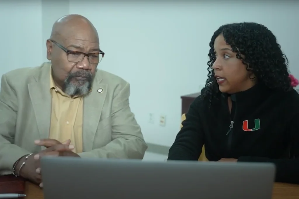 Two individuals seated at a table, discussing something while looking at an open laptop. One wears a beige blazer over a yellow shirt, and the other wears a black jacket with an orange and green 'U' logo. A white wall and furniture are visible in the background