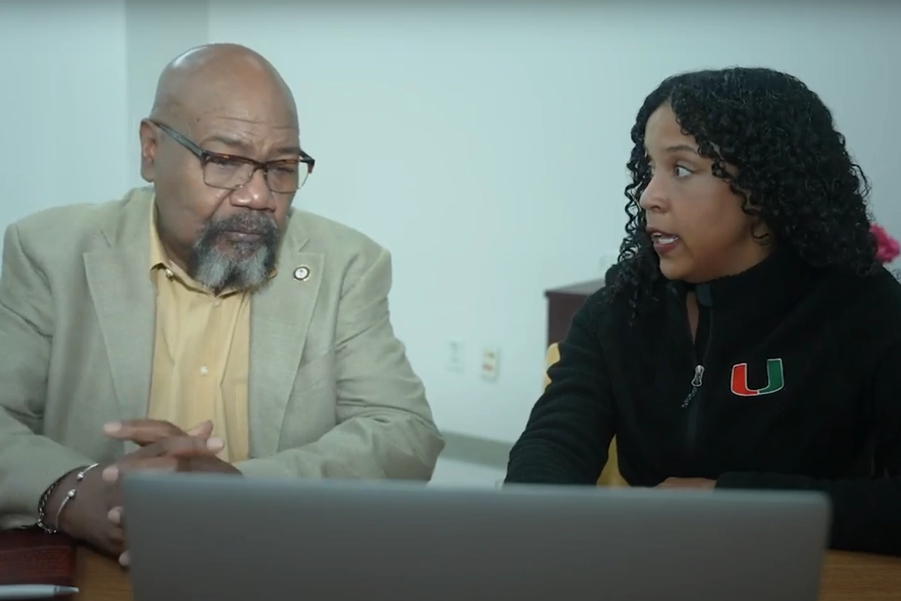 Two individuals seated at a table, discussing something while looking at an open laptop. One wears a beige blazer over a yellow shirt, and the other wears a black jacket with an orange and green 'U' logo. A white wall and furniture are visible in the background