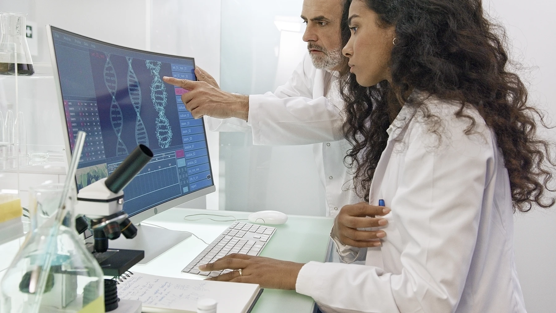 Researchers in white lab coats look at DNA sequencing on a computer screen.