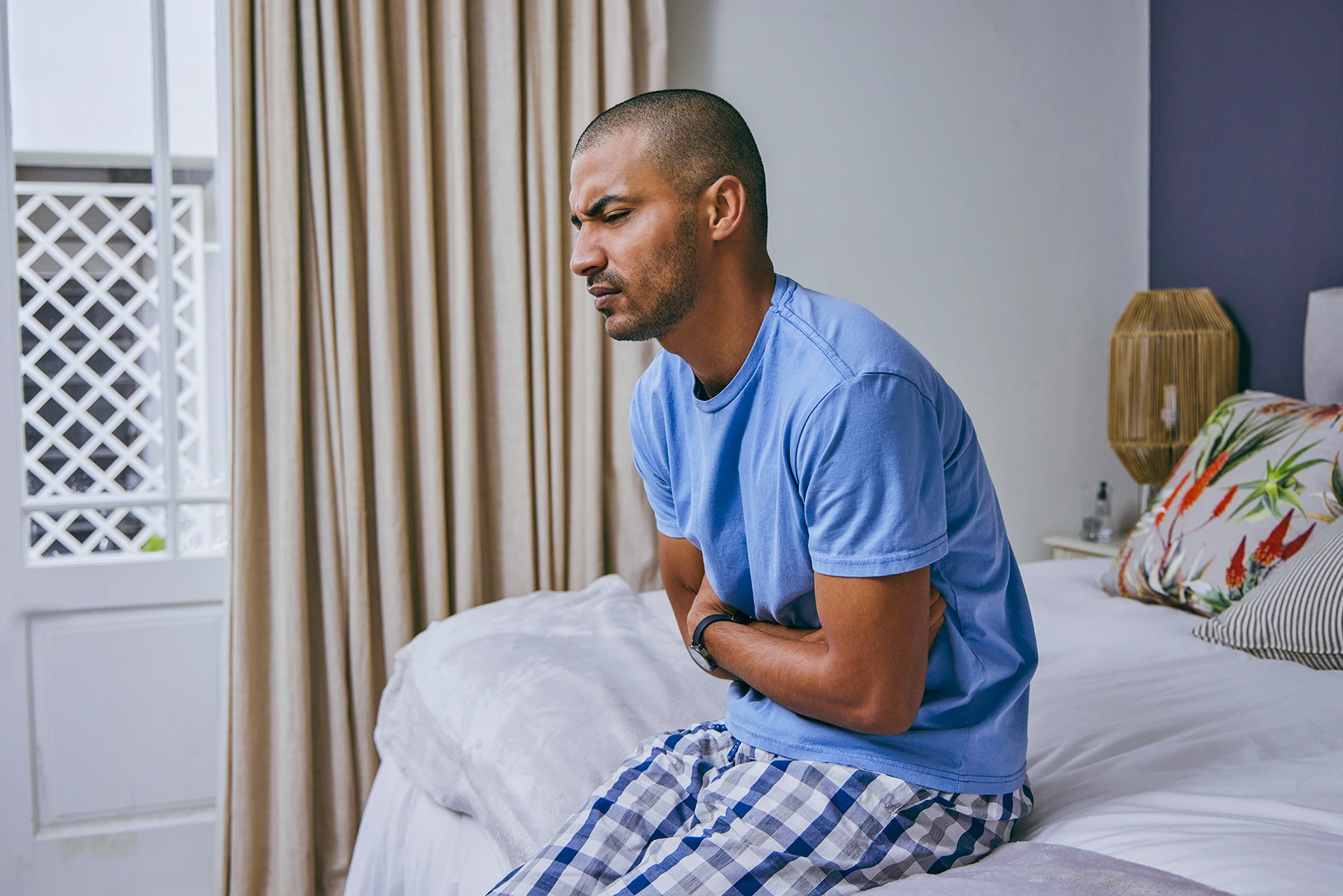 Young Hispanic man sits at the edge of bed, holding his stomach.