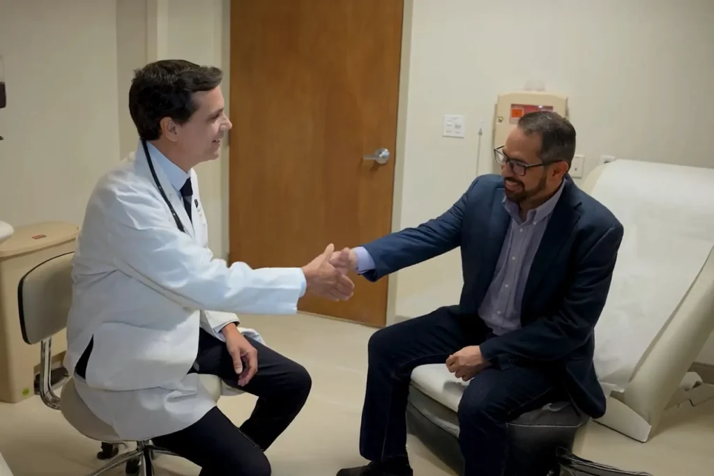 Smiling male doctor in a white lab coat shaking hands with a male patient in a suit jacket, symbolizing a positive healthcare interaction in a medical examination room.