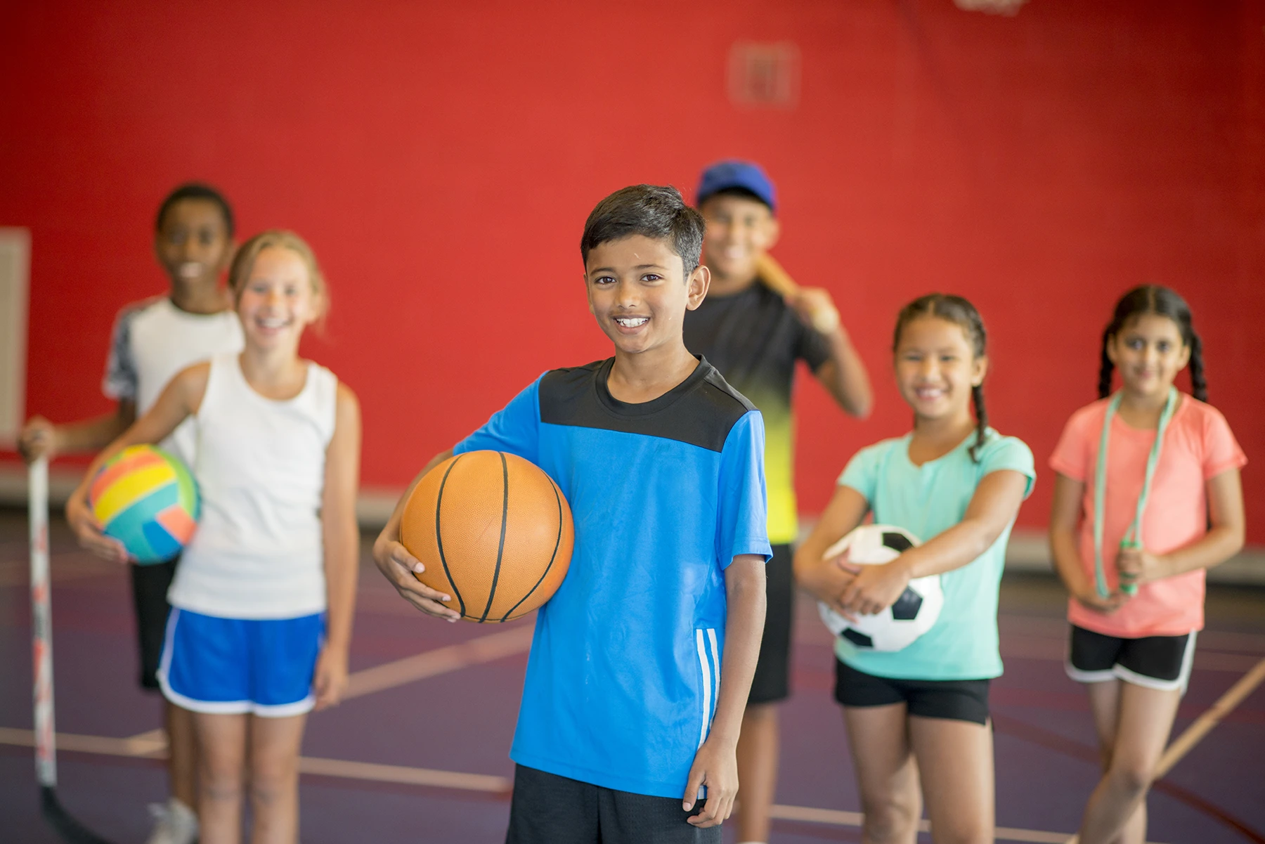 Five kids are standing side-by-side in a gym. They are smiling at the camera and holding sports balls. There is a basketball, soccer ball, two volleyballs, and a dodgeball.