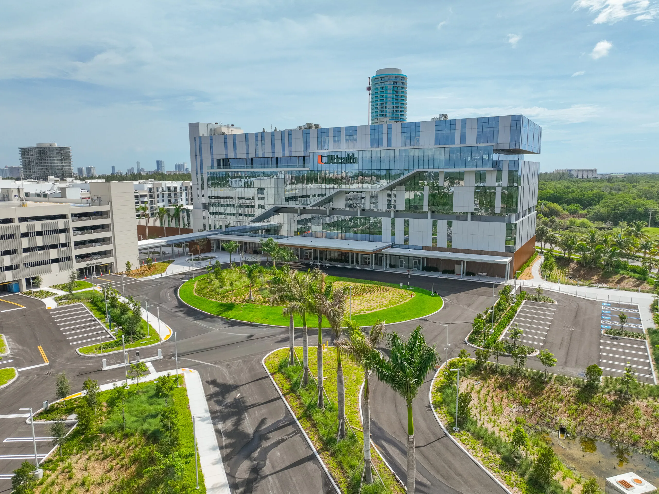 Modern UHealth medical facility featuring a multi-story building with a distinctive glass and white panel facade, surrounded by new parking lots, a parking garage, and lush palm tree landscaping. A distant city skyline and other high-rise buildings are visible under a light blue sky.