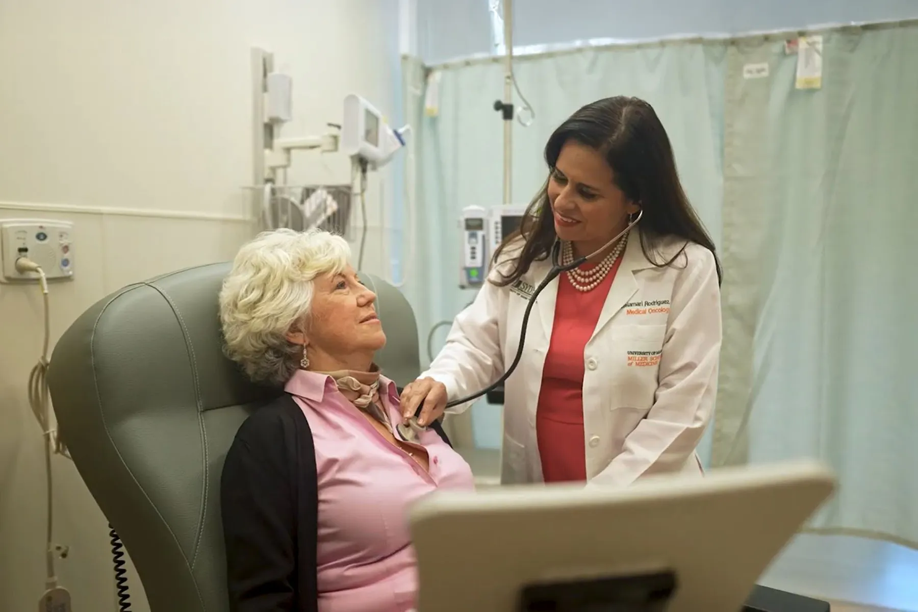 A smiling female doctor in a white lab coat, identified as a Medical Oncologist, uses a stethoscope to examine an older female patient sitting in a medical chair.