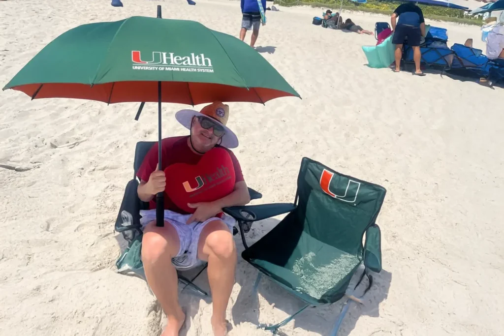 A smiling man on a sunny beach relaxes in a chair under a green UHealth umbrella, holding a red University of Miami heart pillow. An empty matching beach chair is next to him on the white sand.
