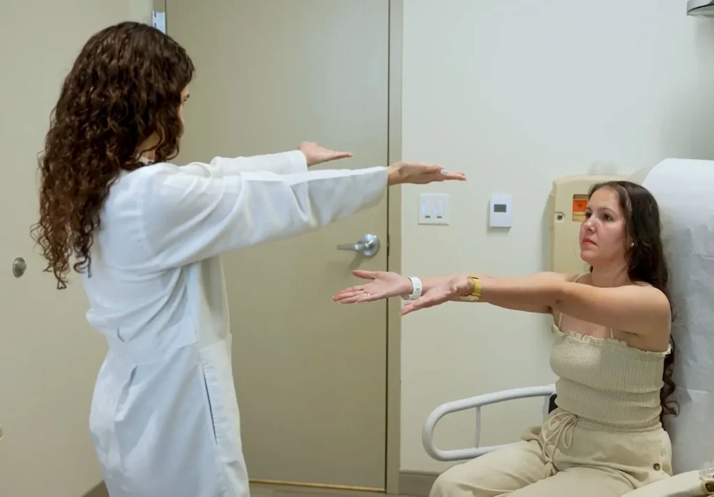 A female doctor in a white lab coat conducts a neurological or balance test with a seated female patient, both holding their arms extended forward with palms facing up in an examination room.