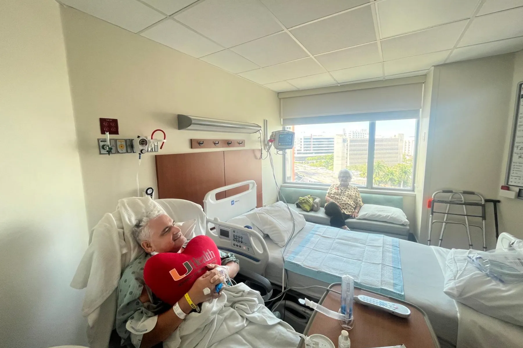 A smiling patient in a hospital bed holds a red UHealth heart pillow, while an older woman sits by a sunlit window overlooking a city skyline in a modern hospital room.