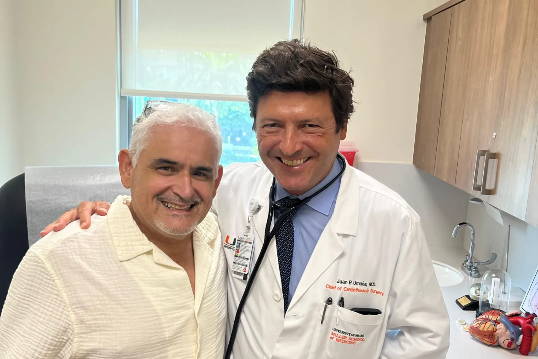 Cardiothoracic surgeon Dr. Juan P. Umaña, from the University of Miami Miller School of Medicine, smiles warmly alongside a patient in a medical examination room. Dr. Umaña wears a white coat and stethoscope, and a heart model is visible on the counter.