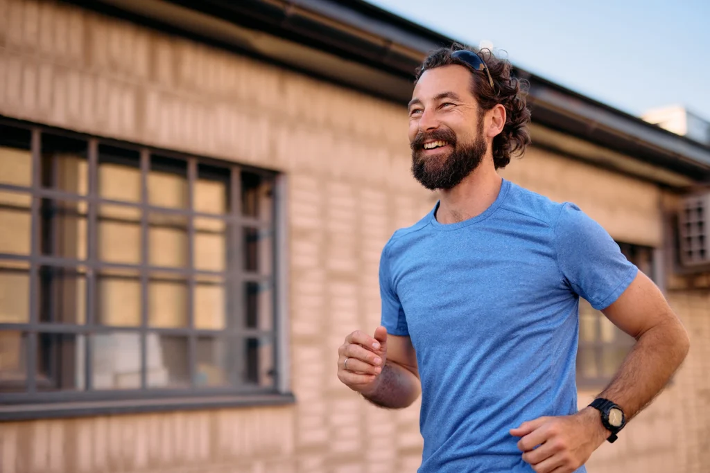 Mid adult man with a beard running energetically in an urban environment, wearing a blue t-shirt, sunglasses, and a wristwatch, smiling while looking away with a sense of joy