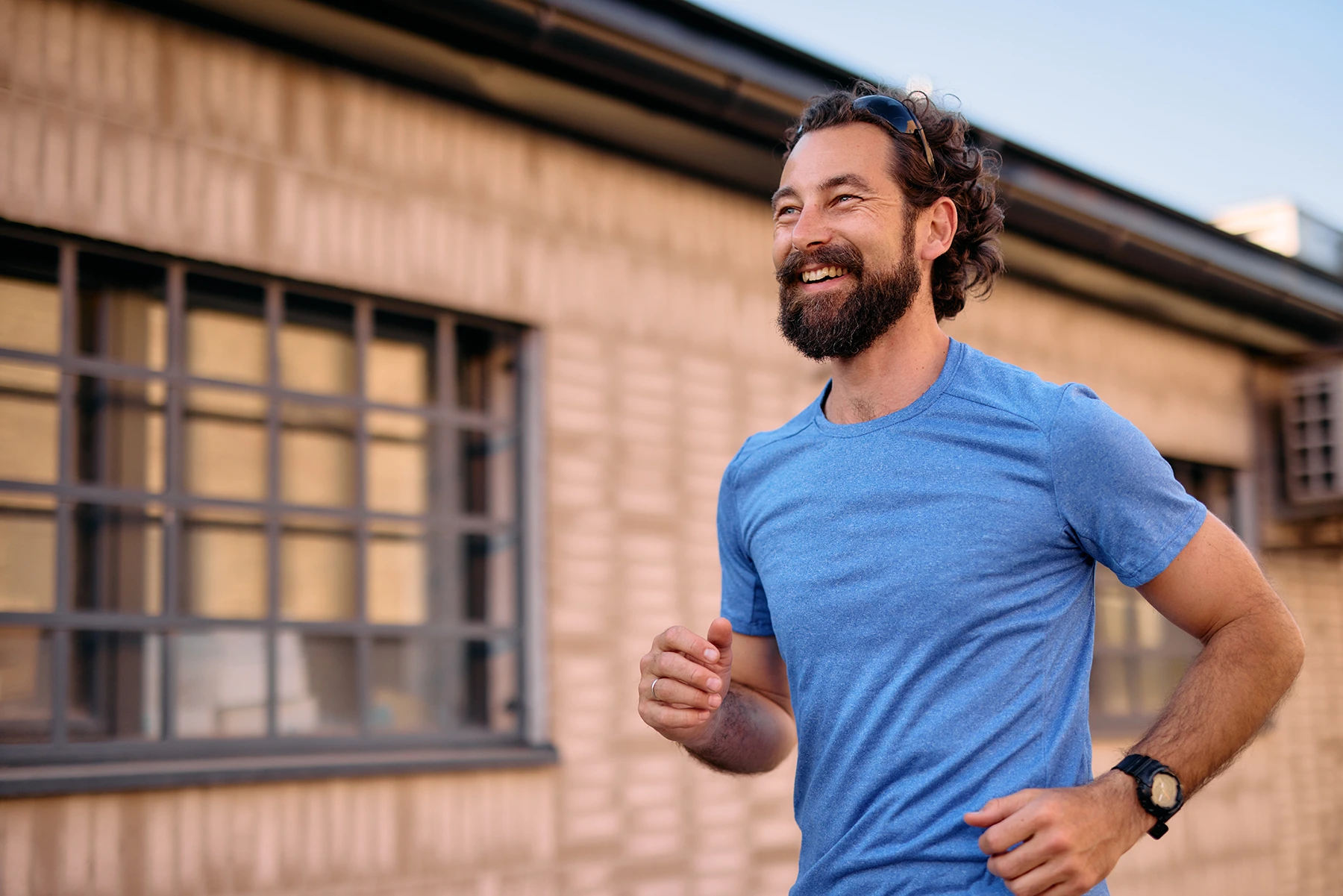 Mid adult man with a beard running energetically in an urban environment, wearing a blue t-shirt, sunglasses, and a wristwatch, smiling while looking away with a sense of joy