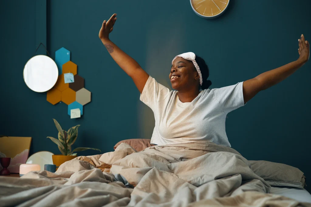 Woman smiling while performing morning stretch in cozy bedroom surrounded by warm decor and green walls with hexagonal wall art creating a relaxing atmosphere