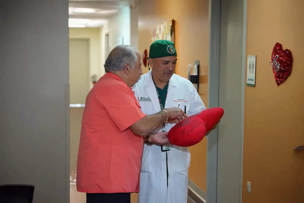 A patient in an orange shirt points at a signed red heart pillow held by Dr. Jorge Reus, a UHealth vascular surgeon, in a hospital hallway decorated with a red tinsel heart.