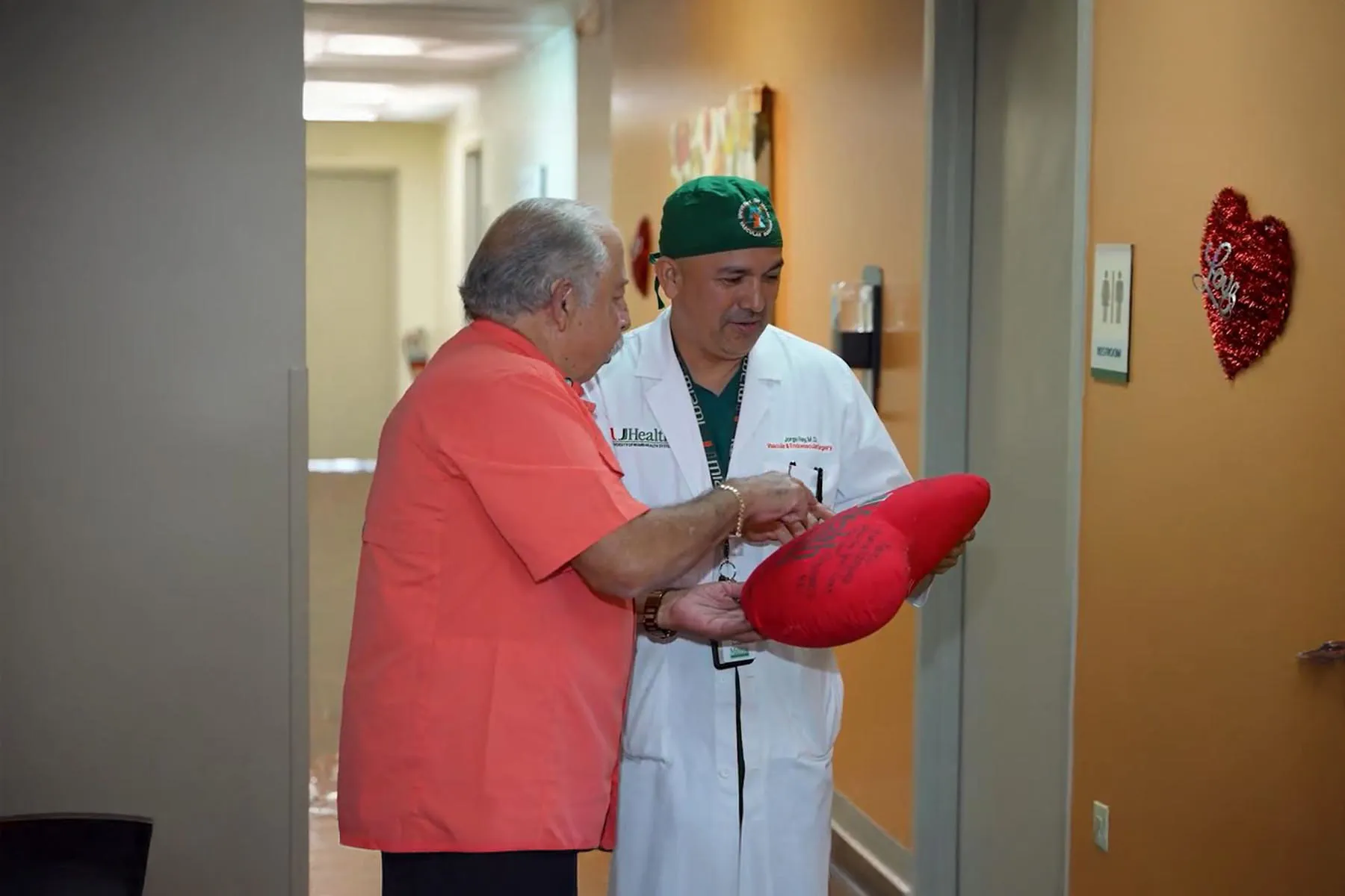 A patient in an orange shirt points at a signed red heart pillow held by Dr. Jorge Reus, a UHealth vascular surgeon, in a hospital hallway decorated with a red tinsel heart.