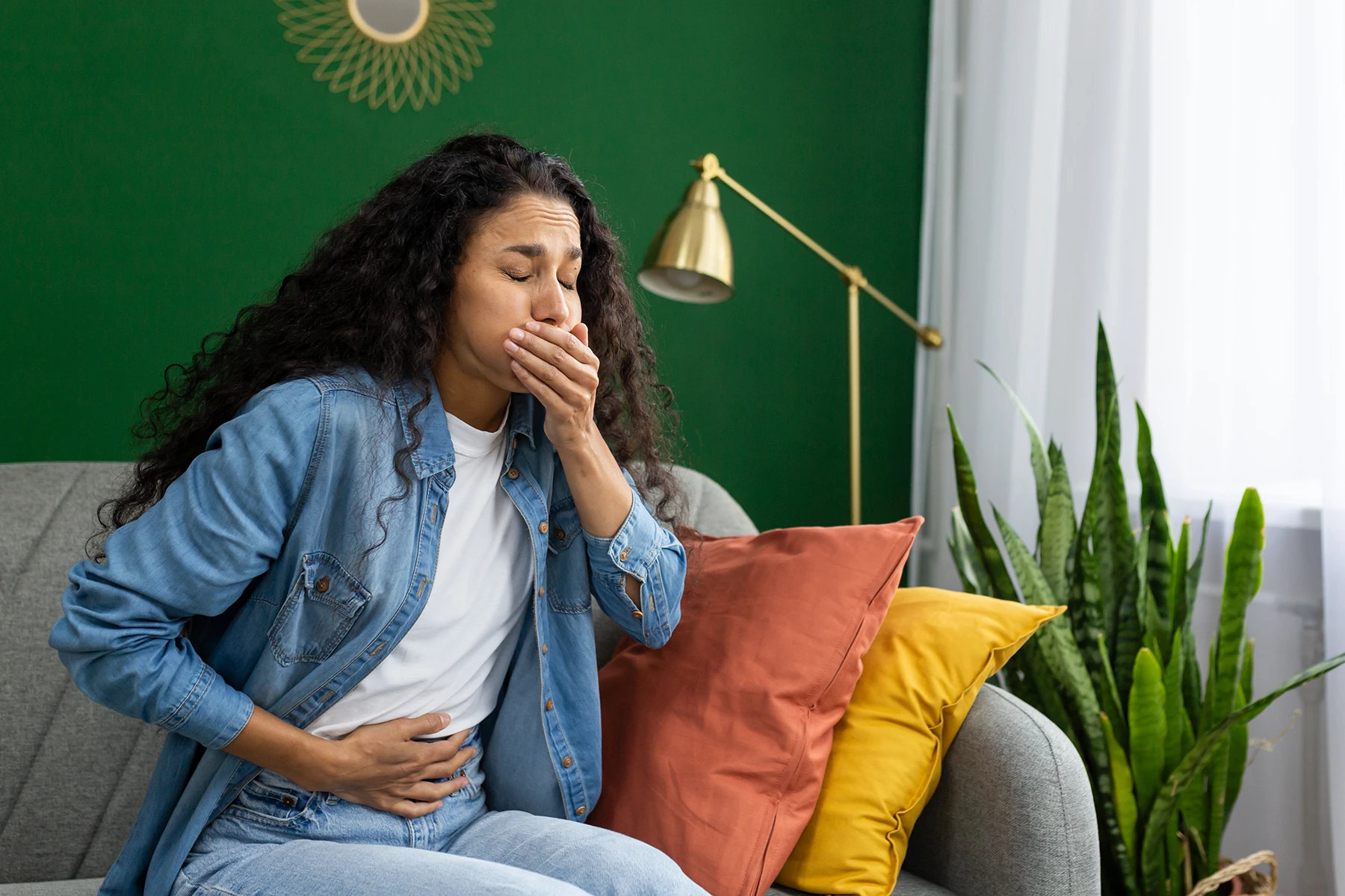 A distressed young woman feeling nauseous on a couch, hand over her mouth, trying to suppress the urge to vomit - stock photo