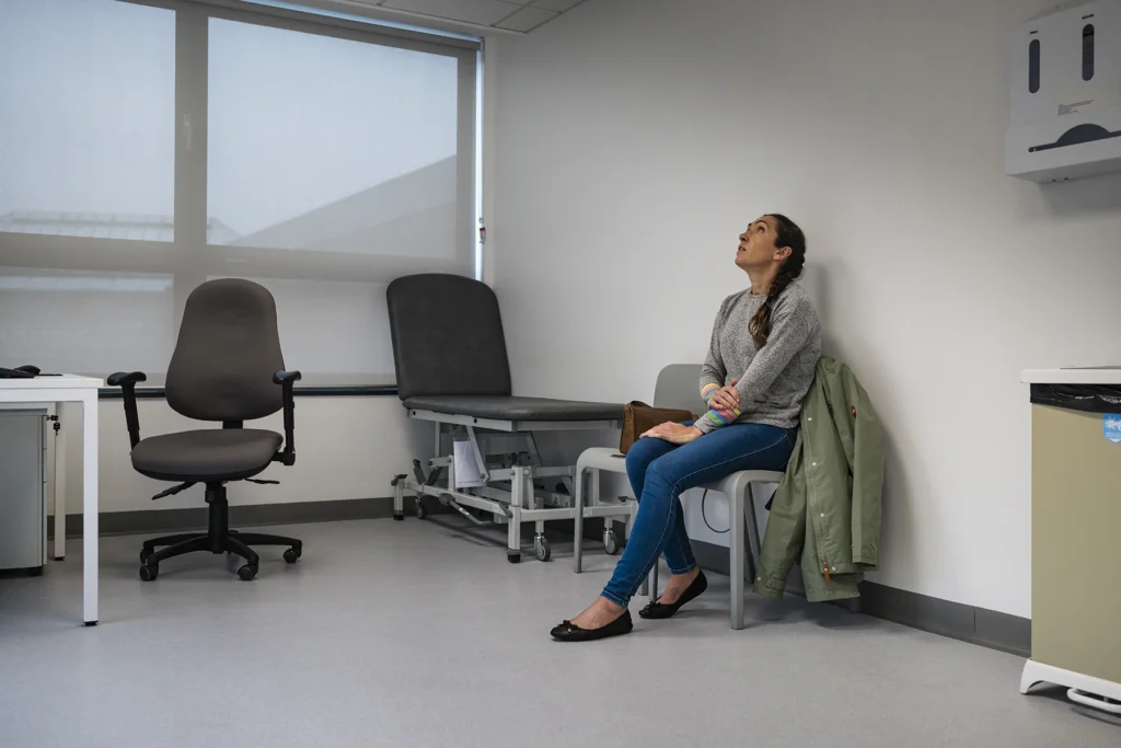 Full length wide view of a mid adult woman sitting in a doctors examination room waiting to be seen, she is holding her arm and looking up, feeling stress. Stock photo