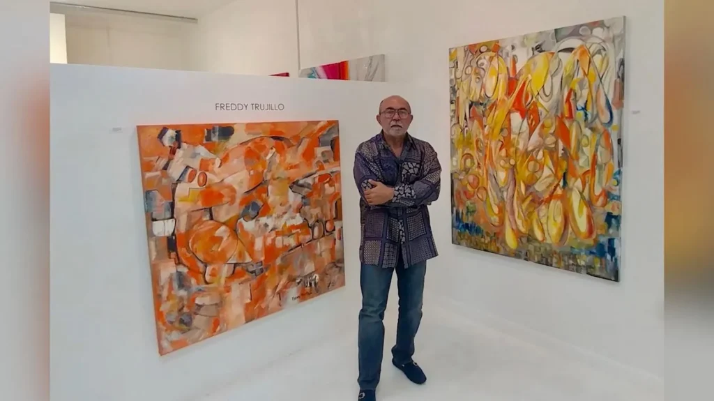 Artist Freddy Trujillo stands in an art gallery, arms crossed, wearing a patterned shirt and glasses, flanked by two large, vibrant abstract paintings. The painting on the left features orange and white tones, while the one on the right is dominated by yellows and oranges with flowing shapes.