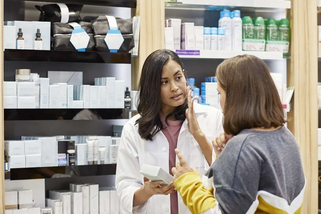 Woman explaining while selling skin care product. Customer is listening to pharmacist standing against rack. They are discussing at pharmacy.