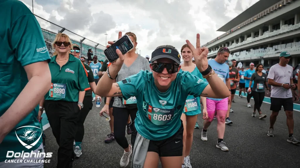 Wearing sunglasses and Dolphins Cancer Challenge running shirt, Claudia Pereira smiles and flashes a peace sign while walking with a group of race participants.