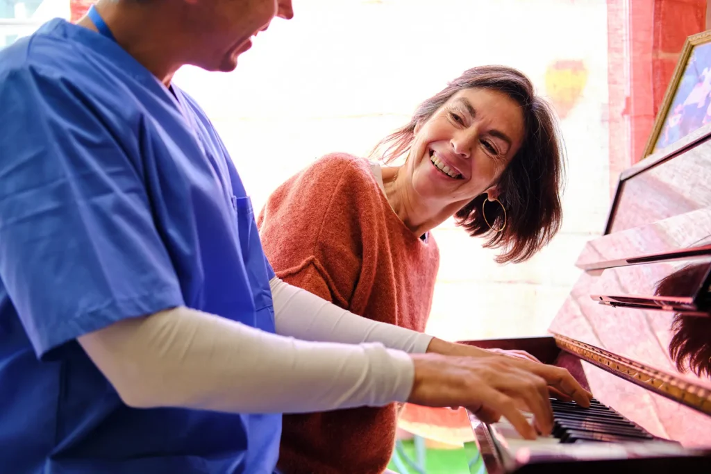 Nurse playing piano with laughing senior adult woman, providing music therapy.