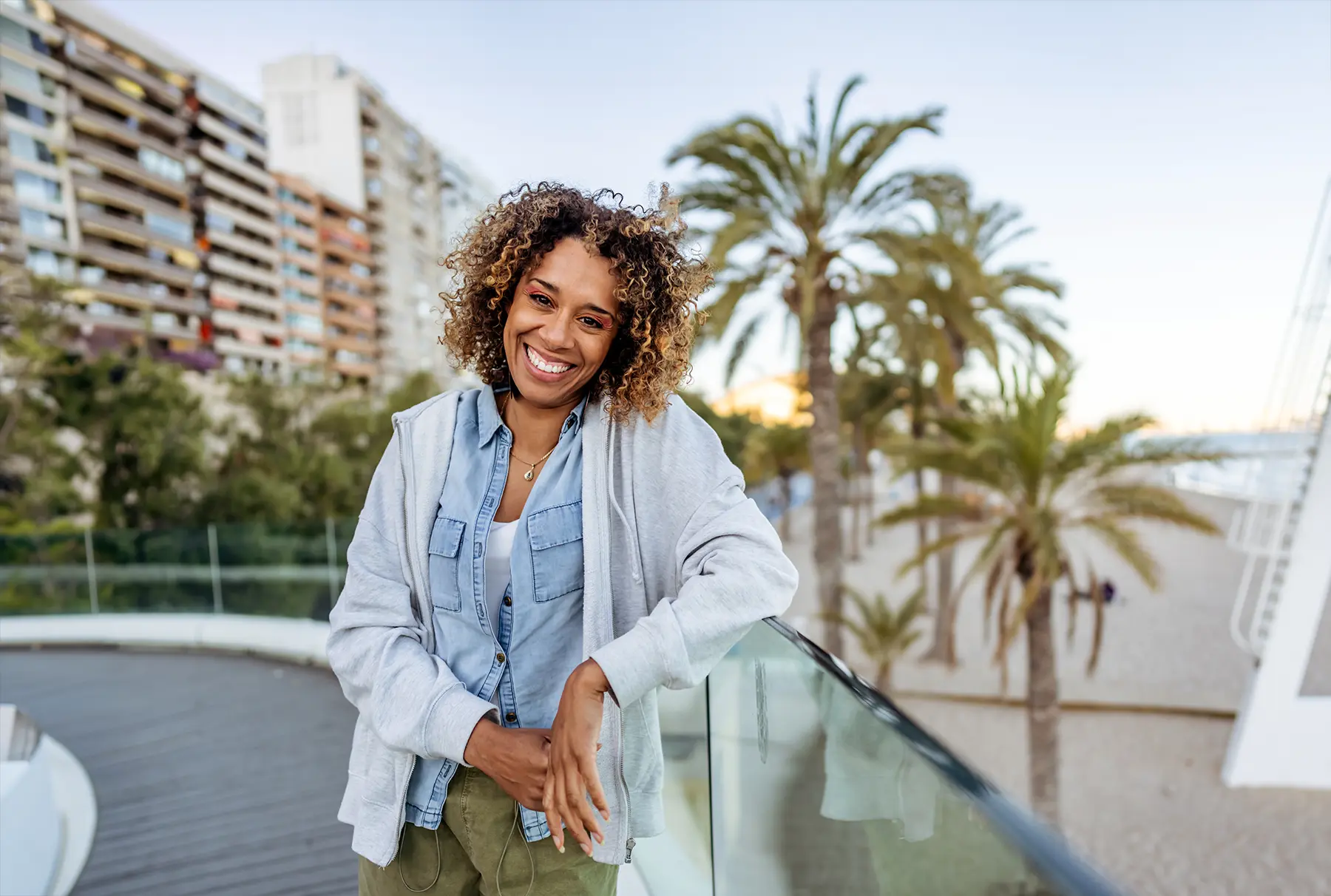 Cheerful woman in a casual outfit standing on a boardwalk near palm trees and the beach, radiating positivity and enjoying a sunny day. Urban buildings and greenery create a harmonious urban-beach combination.