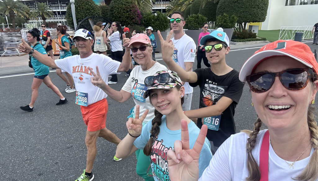 Susan Clark participates in the Dolphins Cancer Challenge alongside family members and fellow participants on a roadway lined with event attendees. Several participants wear race bibs and athletic apparel while making peace signs during the walk. Palm trees, landscaping and event activity are visible in the background.