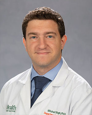 Professional headshot of University of Miami Health System physician Dr. Mitchell Slugh wearing a white medical coat, light blue dress shirt and dark blue tie against a neutral gray studio background. The physician is facing the camera in a chest-up portrait with even lighting and a clean, clinical appearance.