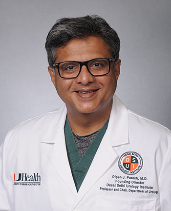 Professional portrait of Dr. Dipen Parekh in a white medical coat and surgical scrubs against a neutral gray background. The physician wears glasses and is pictured from the chest up in a studio-style headshot used for academic medicine and clinical leadership profiles.