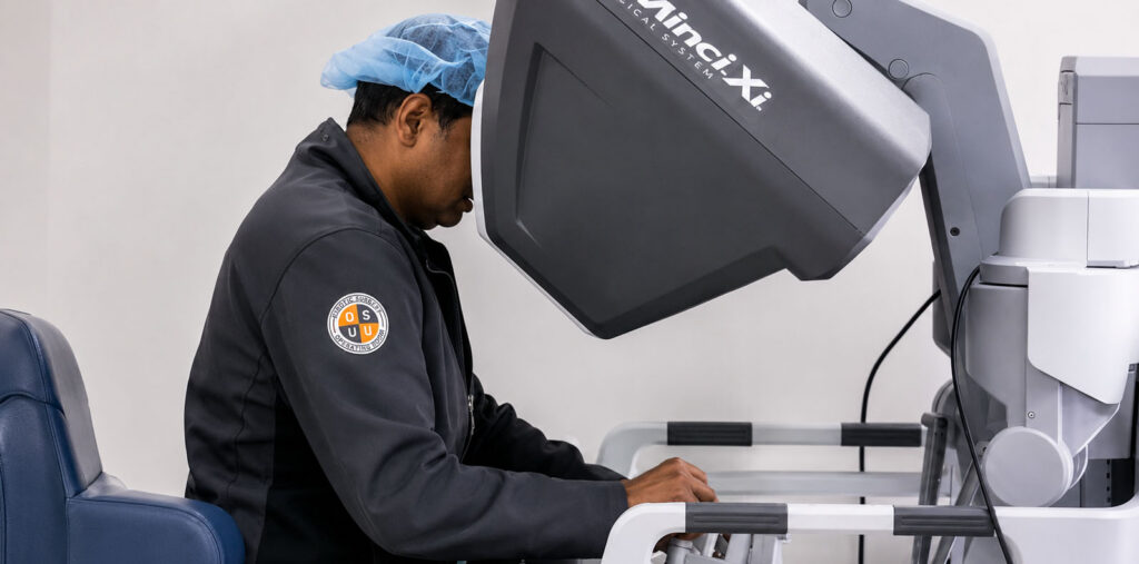 A surgeon seated at a robotic surgical console operates a da Vinci surgical system in a clinical setting. The individual wears a surgical cap and scrubs while using hand controls and a viewing interface that enables minimally invasive robotic-assisted procedures.