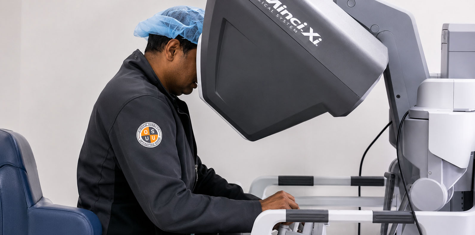 A surgeon seated at a robotic surgical console operates a da Vinci surgical system in a clinical setting. The individual wears a surgical cap and scrubs while using hand controls and a viewing interface that enables minimally invasive robotic-assisted procedures.