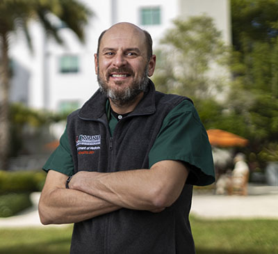 Dr. Justin Watts standing outside of Sylvester Comprehensive Cancer Center, wearing a green shirt and black pullover, his arms crossed over his chest