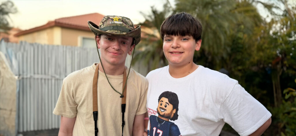 Jake and Justin Kleppen stand side by side outdoors in a residential yard. One wears a camouflage U.S. Army-style hat and a light-colored shirt with binocular straps around the neck. The other wears a white graphic T-shirt. A fence, palm trees, and a house are visible in the softly blurred background.