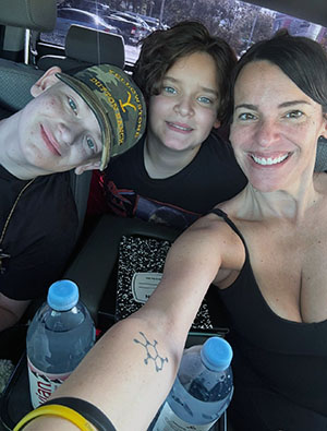 Justin, Jake and Marisel Teppen pose together inside a vehicle for a selfie. Two individuals sit in the back seat while one extends an arm from the front seat to take the photo. Water bottles and notebooks are visible in the foreground, with parked vehicles seen through the windows in the background.