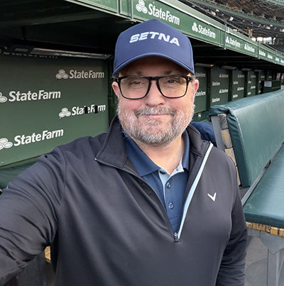 Ryan wearing a navy baseball cap, glasses, and a dark quarter-zip jacket takes a selfie from a baseball stadium dugout. Green seats and stadium signage are visible in the background, with the word “StateFarm” appearing on multiple signs behind the seating area.