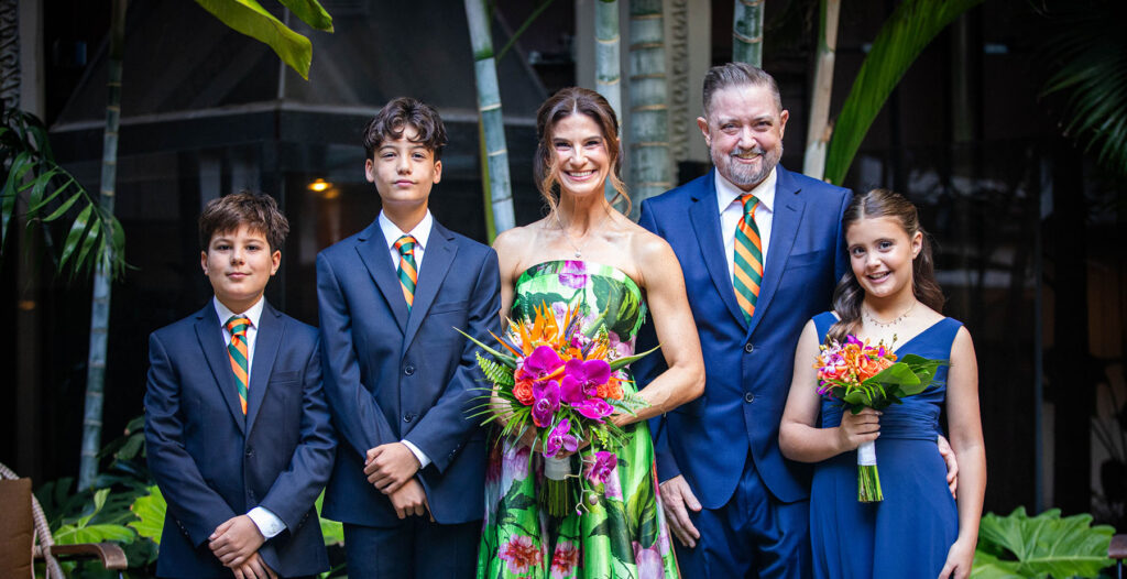 Ryan Goepel and family standing together in a lush indoor setting surrounded by tropical plants. Two adults stand in the center, flanked by three children. The adults and two children wear coordinated blue formal attire with orange-and-green striped ties, while one child wears a blue dress and holds a bouquet. The adult in the center and the child in the blue dress also hold colorful tropical flower bouquets. The group faces the camera in front of bamboo and greenery.