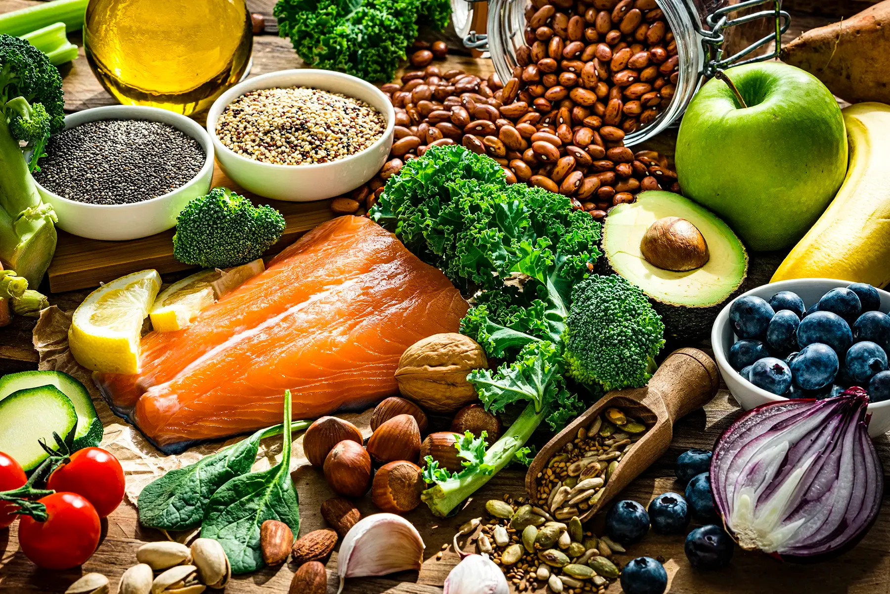 Variety of healthy and nutritious whole foods, including fresh salmon, avocado, broccoli, kale, blueberries, quinoa, nuts, and olive oil, arranged on a rustic wooden table.