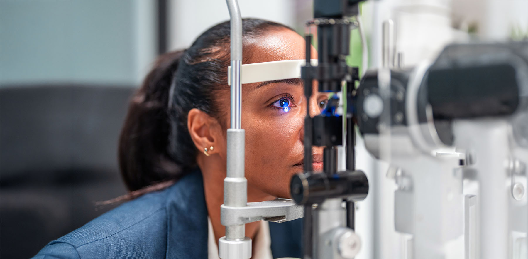 Hispanic female patient positioned at slit lamp apparatus for eye examination. Optometrist conducting diagnostic assessment of eye health and vision. Clinical setting with specialized optical equipment and professional lighting.