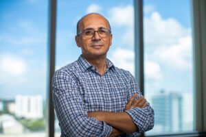 Dr. Michael Benatar stands with arms crossed in front of large office windows overlooking the Miami skyline.