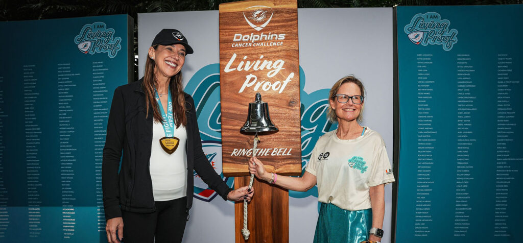 Jennifer Buttrick rings the bell at a Dolphins Cancer Challenge event while standing beside Dr. Carmen Calfa in front of “Living Proof” displays honoring cancer survivors.