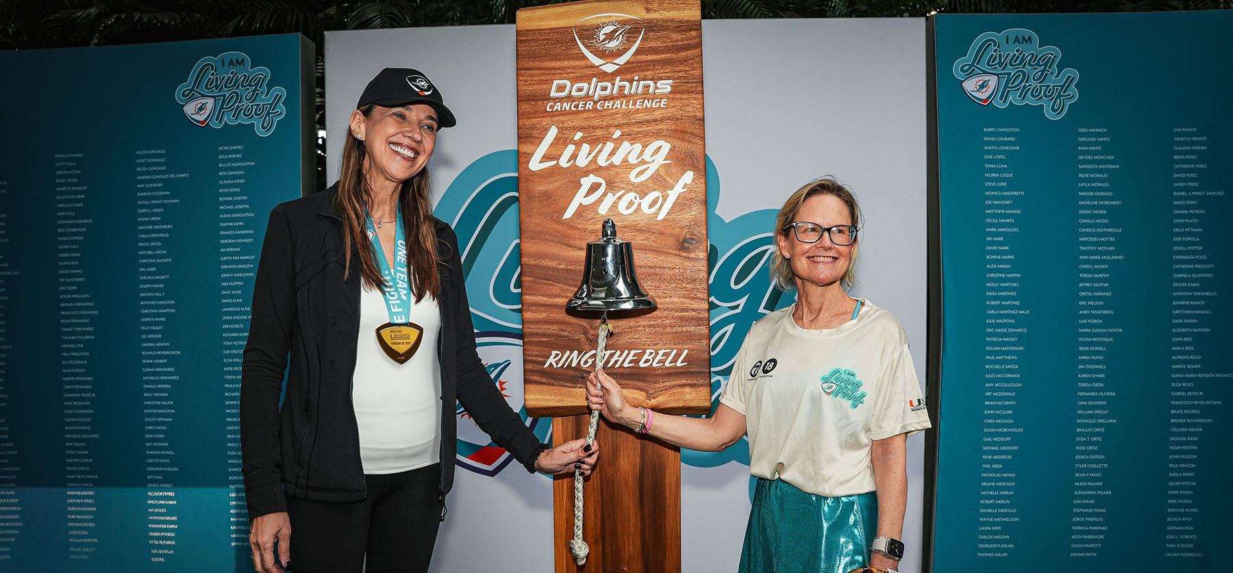Jennifer Buttrick rings the bell at a Dolphins Cancer Challenge event while standing beside Dr. Carmen Calfa in front of “Living Proof” displays honoring cancer survivors.